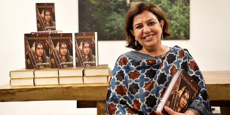 Saba Dewan wearing a blue black and red dupatta holding a copy of her book and standing next to a stack of other copies. She is smiling.