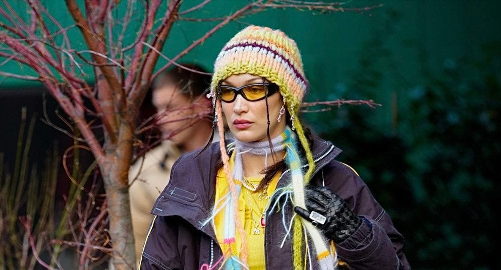 bella hadid wears a multicolored knitted beanie, a weird stringy scarf, and yellow-lens sunglasses with black frames.