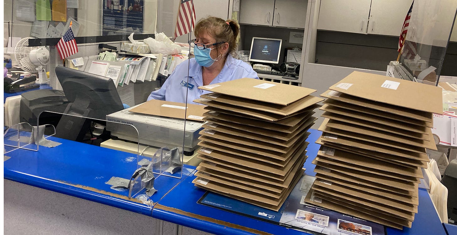 A scene at a post office counter. A woman with brown hair in a pony tail, glasses, and a mask weighs a brown cardboard envelope. There are two tall stacks of envelopes on the counter.