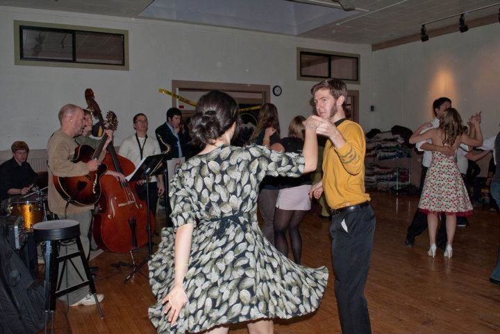 Jessie's back is to the camera as she dances with a male dance partner. Her dress patterned with green leaves is fanned out in a twirl. A small band plays in the background as other dancers dance.