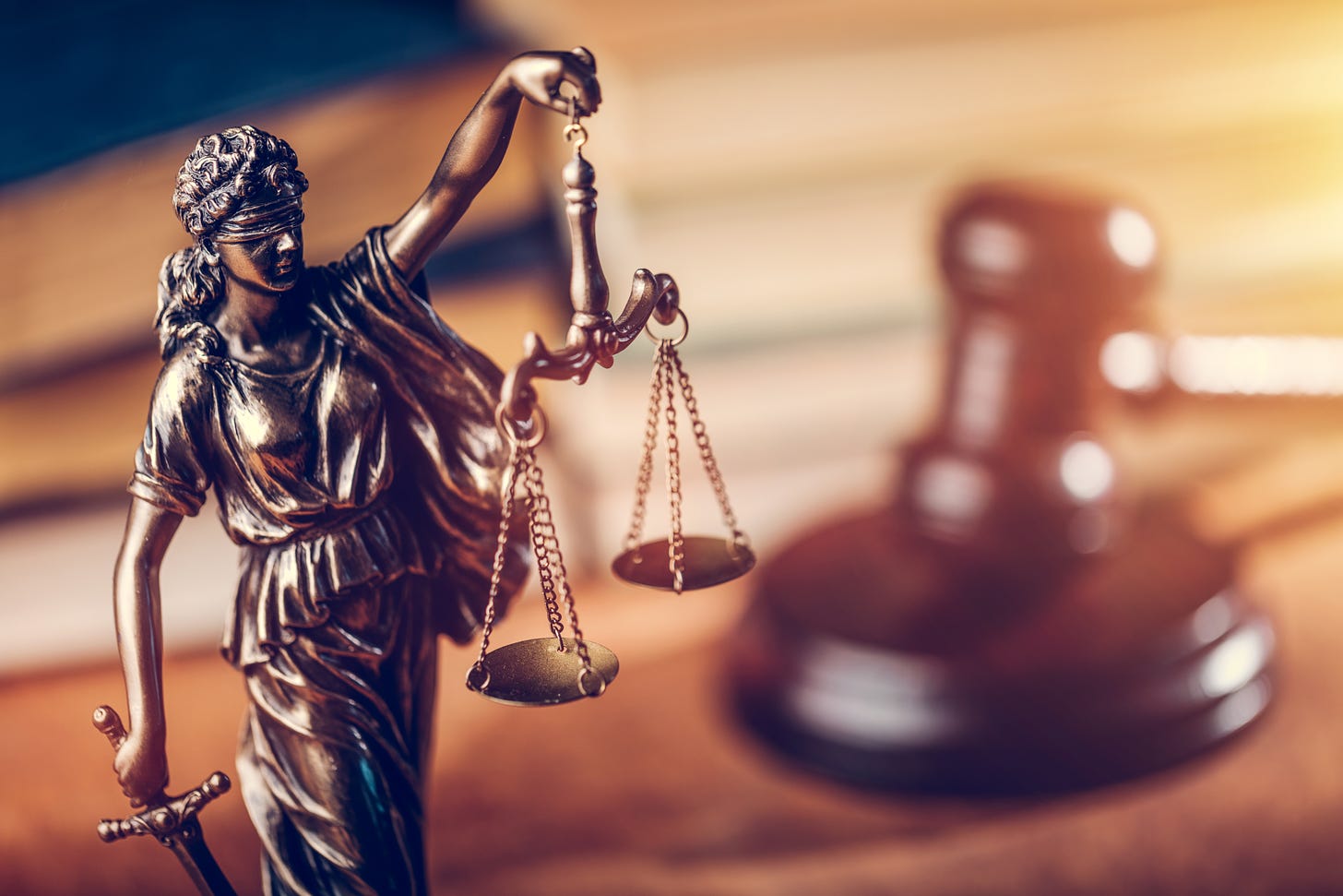 Image contains Lady Justice/Themis statue in the foreground, made of bronze.  She's holding up the scales of justice in her left hand, and her right hand is at her side, holding her sword.  The background is blurred and contains a judge's gavel and old law books with black covers.