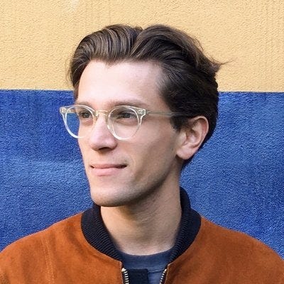 Foto de Dan Abramov, em 3x4, de óculos, jaqueta, olhando à esquerda, levemente sorrindo, com uma parede de fundo.