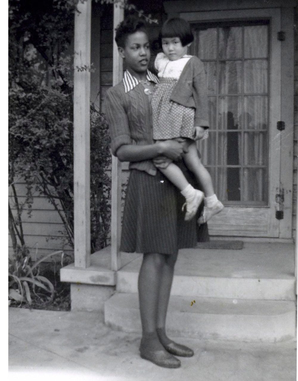 Archival black and white image of a Black woman holding a Japanese American child on the porch of a house.