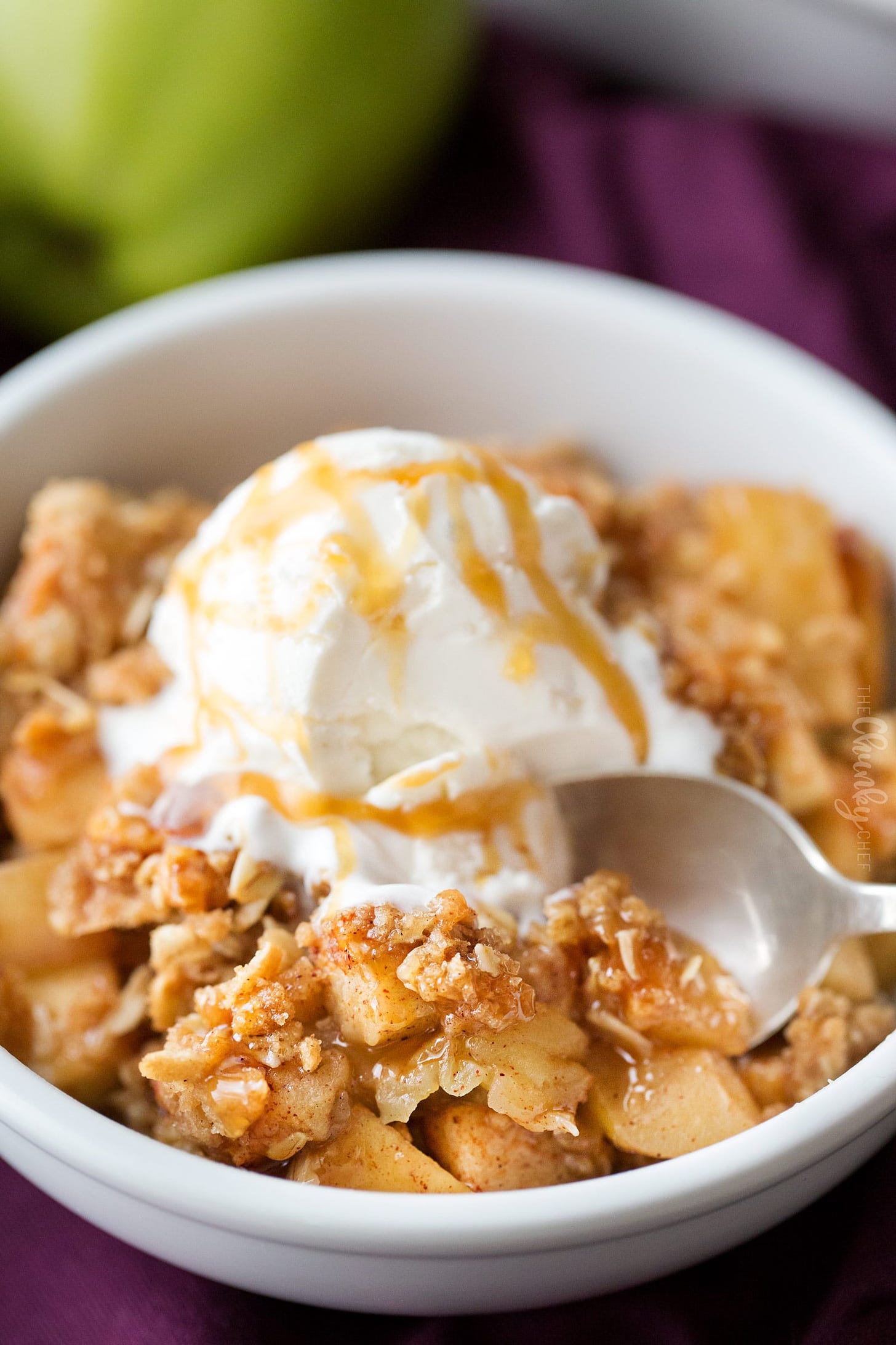 Apple crisp in a bowl with scoop of vanilla ice cream and caramel sauce, with a spoon taking a scoop