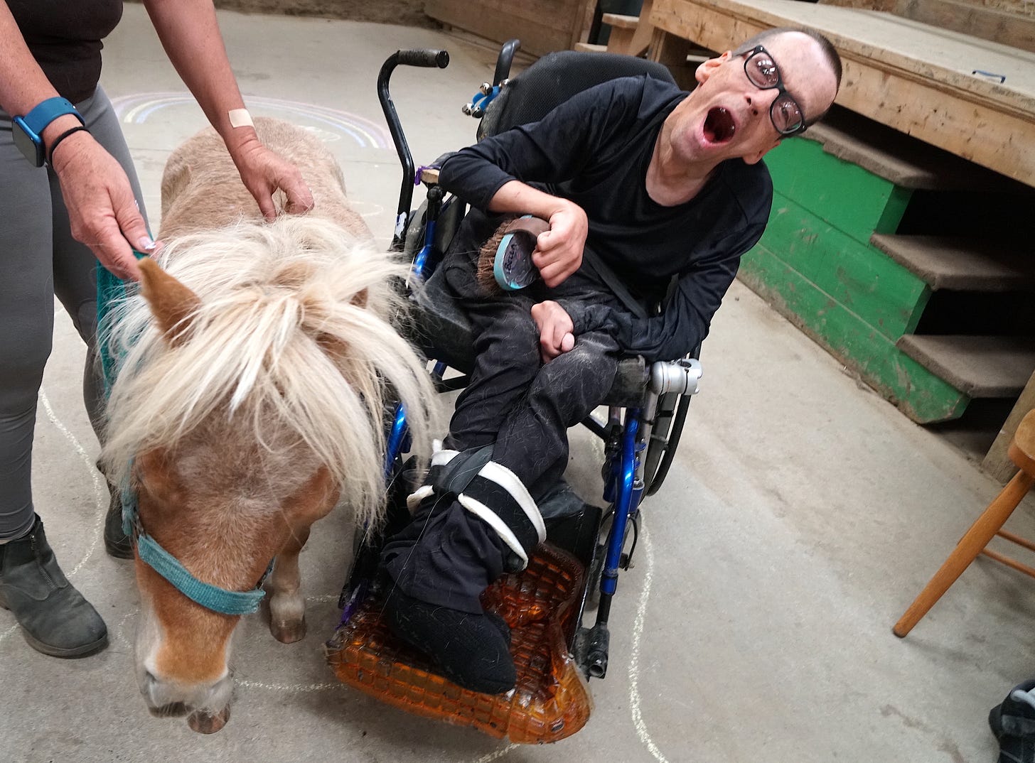 A man with a crew cut, wearing dark-colored clothing and dark-framed glasses, leans to the side in his wheelchair, his gap-toothed mouth opened widely. He holds a grooming brush in his contorted right hand as a miniature horse stands beside him.