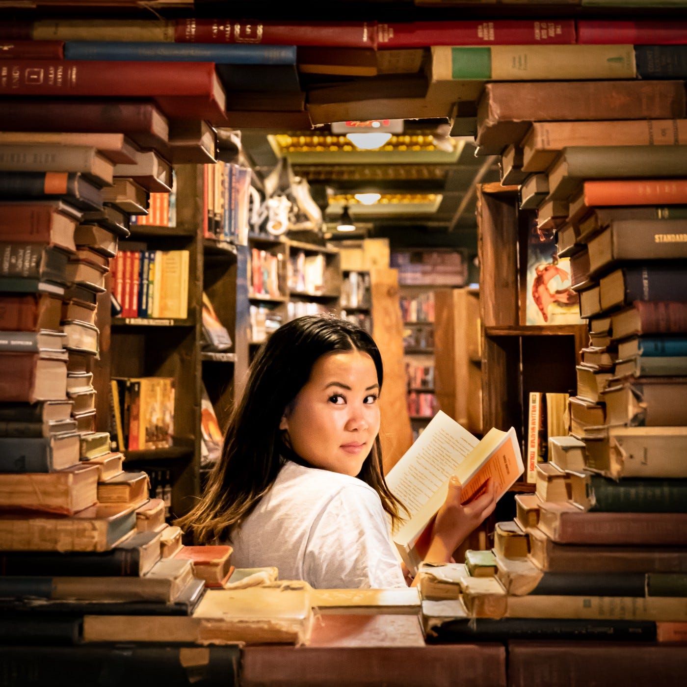 A woman reading a single paper book in the middle of piled books and books on shelves. She looks back at the viewer with a smile.