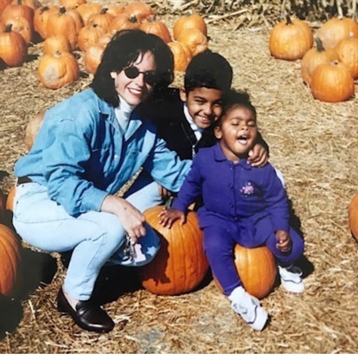 family of three in a pumpkin patch, mom to left in denim shirt, little boy in middle, and little girl to left sticking her tongue out