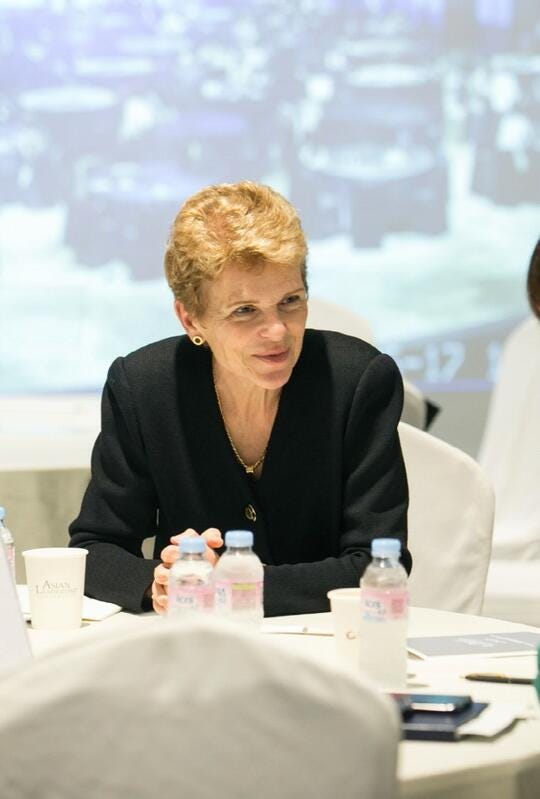 A white woman with short blond hair sits at a table. She is wearing a dark suit which contrasts against the white tablecloth and light colored background. 
