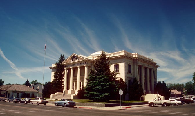 Photograph of the Elko County Courthouse in 1989 with a blue sky above . The courthouse was built in 1911 and has typical features of the period, with Greek column entrances on three sides and a dome on top.