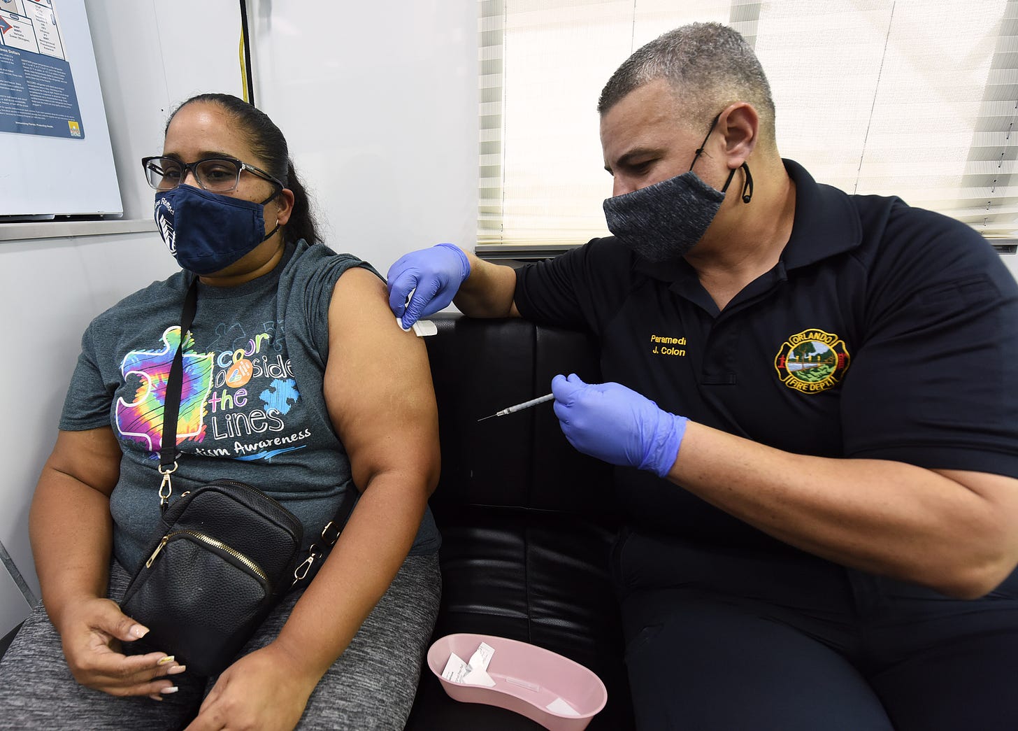 A man in an Orlando Fire Department polo holds a syringe and dabs the arm of a woman in a mask with alcohol, ready to administer the vaccine.