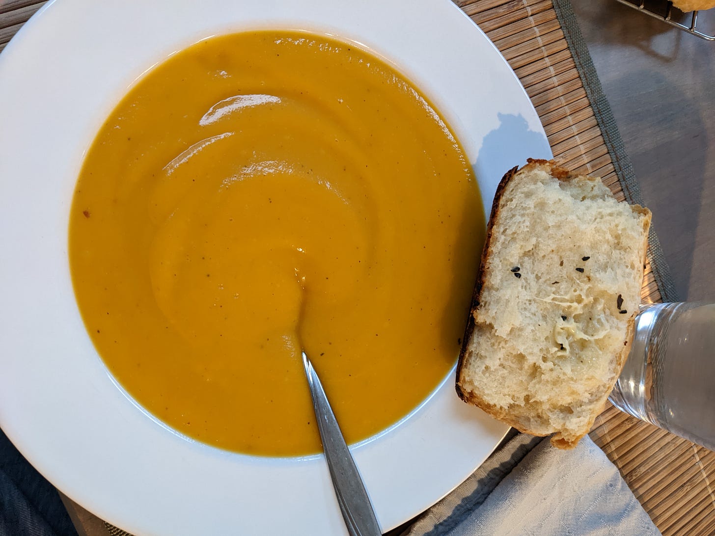 A bowl of butternut squash soup in a white bowl with a piece of bread on the right side.