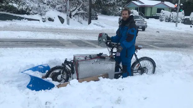 On a snowbound street, a man stands on a stationary cargobike with a small snowplough attached to the front. 
