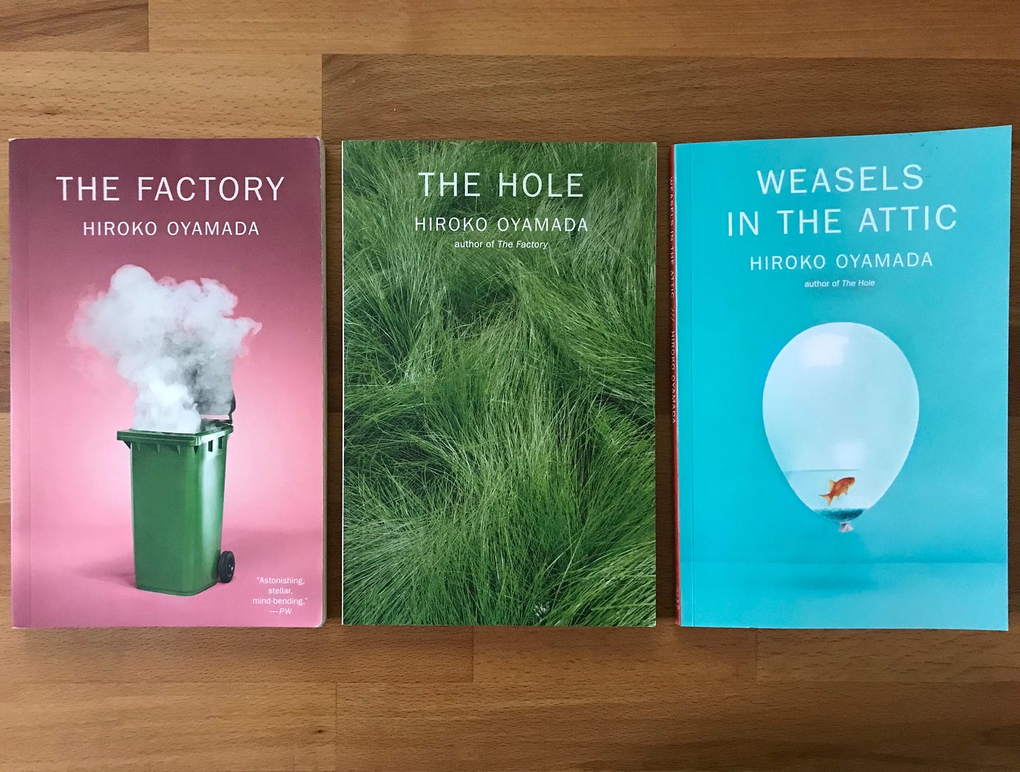 Three books by Hiroko Oyamada laid side-by-side. One has a smoking garbage can, another wavy long grass, and the other a balloon filled with a little bit of water and a goldfish.