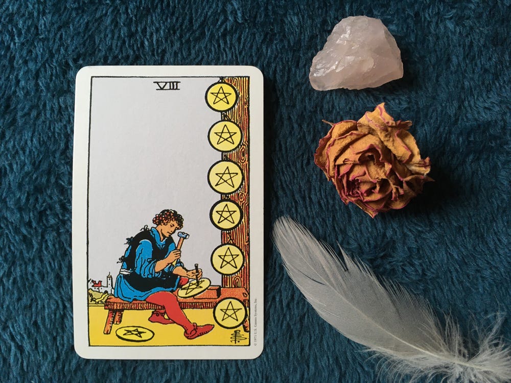 The Eight of Pentacles Tarot card (person carving gold coins) on a blue fleece blanket, next to a rose quartz stone, a dried rose, and a grey feather.