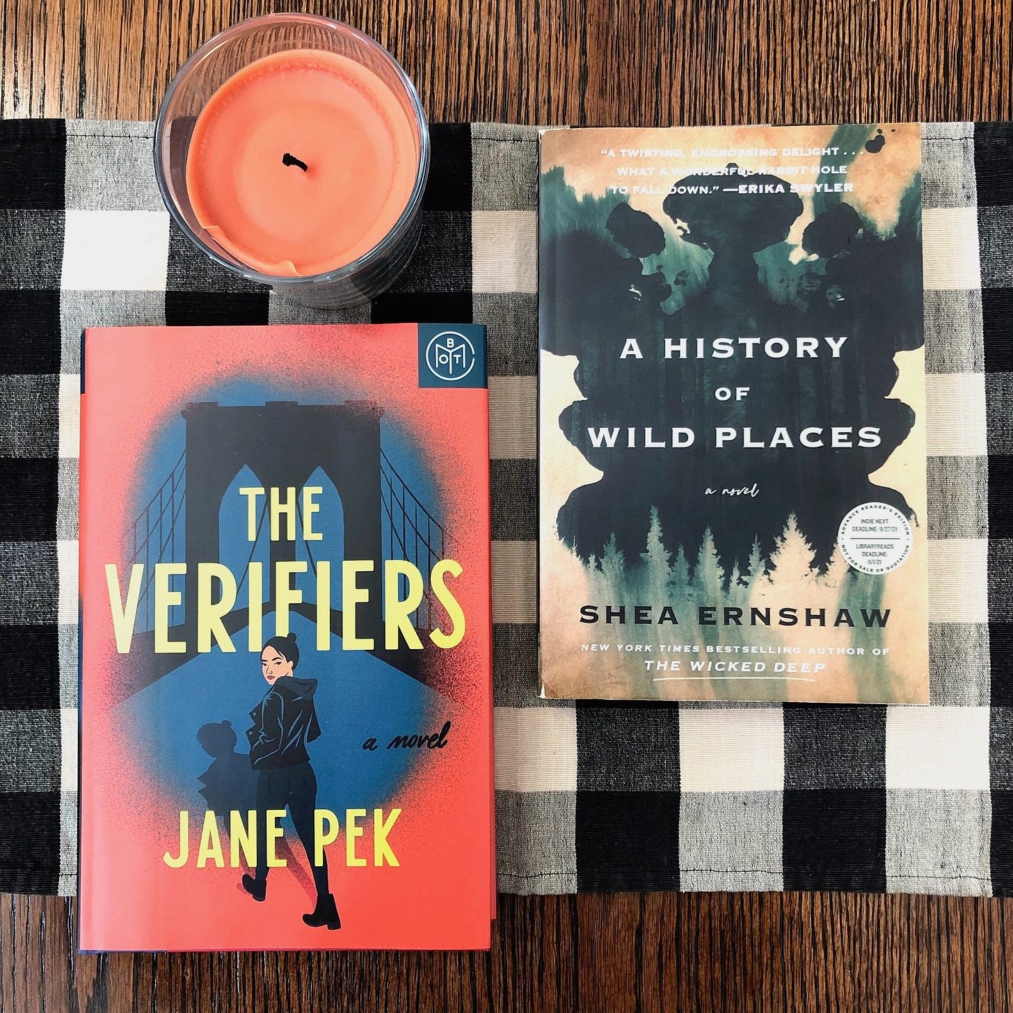 The books The Verifiers and A History of Wild Places, laying on a checked placemat on a wood table next to an orange candle. 