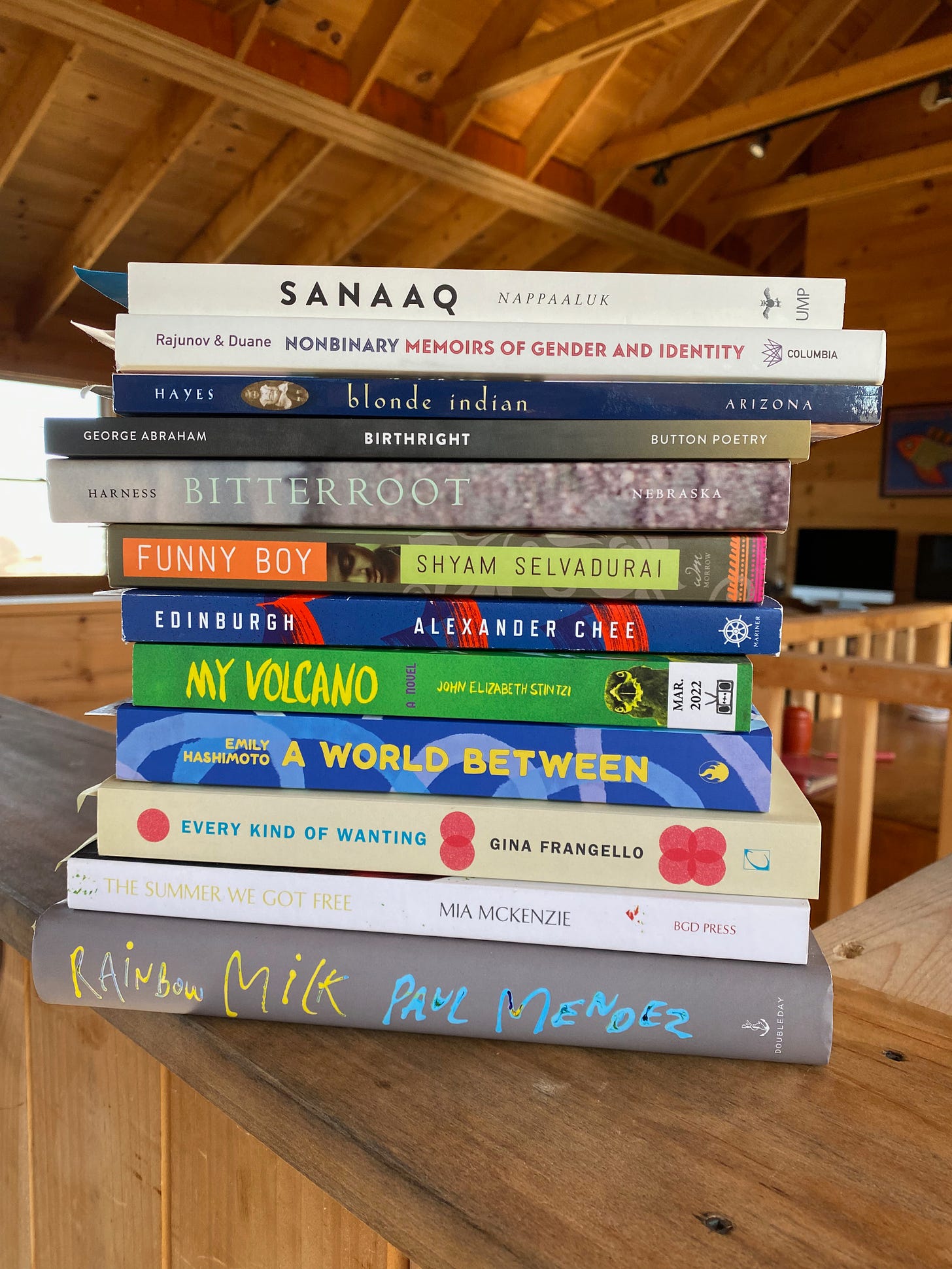 A stack of books on a stair ledge, with wooden roof rafters visible in the background. Books are: Sanaaq, Nonbinary: Memoirs of Gender and Identity, Blonde Indian, Birthright, Bitterroot, Funny Boy, Edinburgh, My Volcano, A World Between, Every Kind of Wanting, The Summer We Got Free, and Rainbow Milk.