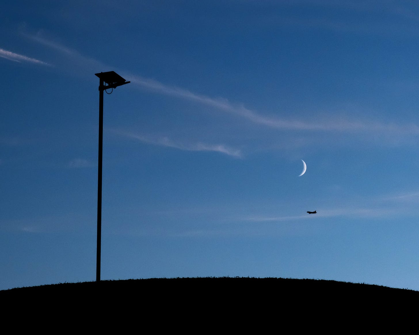 A plane flies under a crescent moon in the distance. The photograph is taken from a hill shown in silhouette. A light post at the top of the hill provides a sense of scale to the scene.
