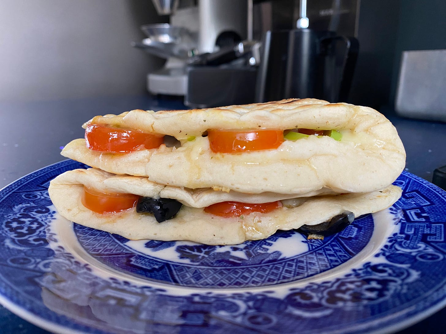 A blue and white plate with a folded flatbread filled with cheese, tomato, black olives and green onion. 