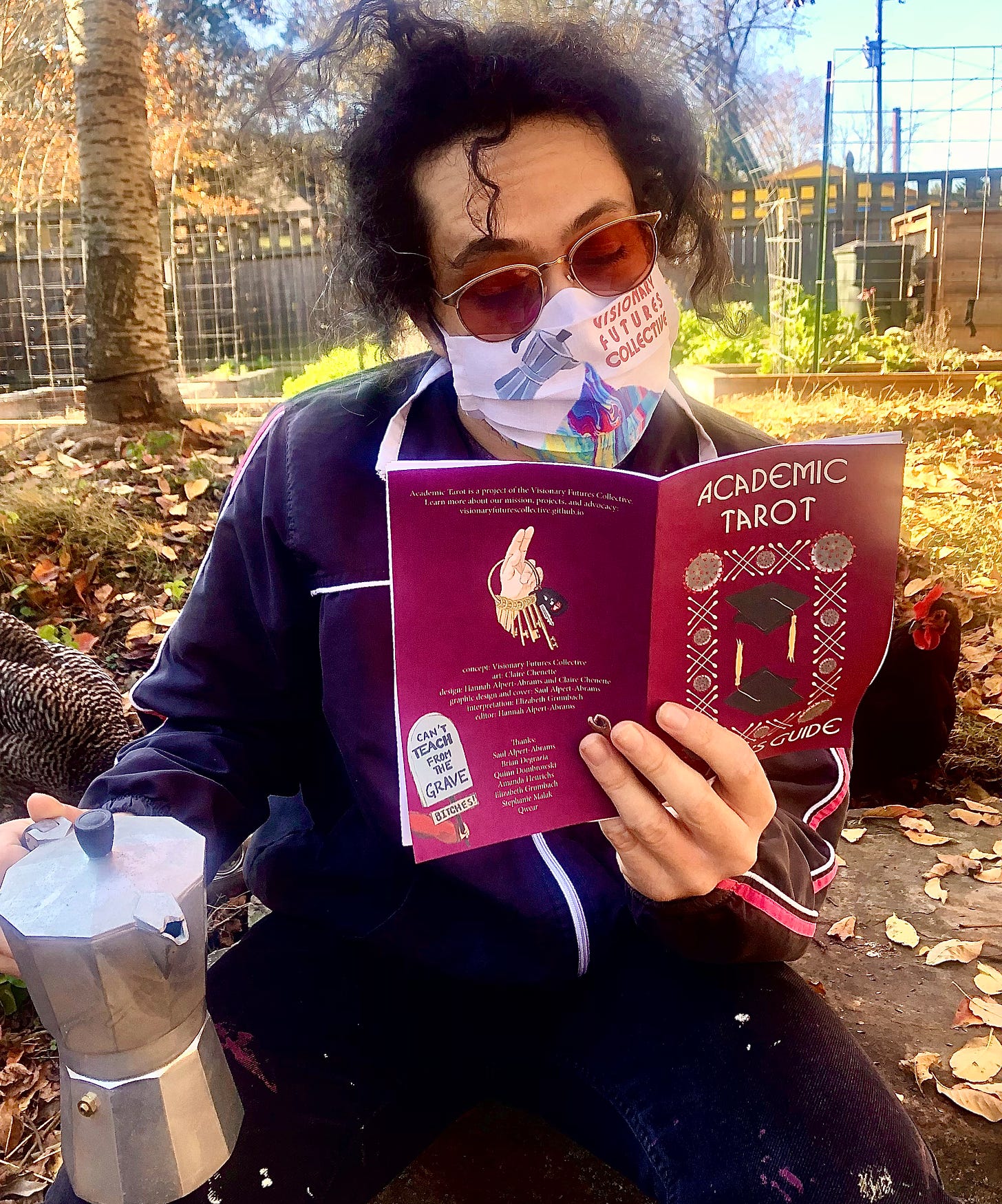 A white man with curly hair holds a Moka pot while reading the Academic Tarot reader's guide in a sunny autumnal courtyard, while chickens look on.