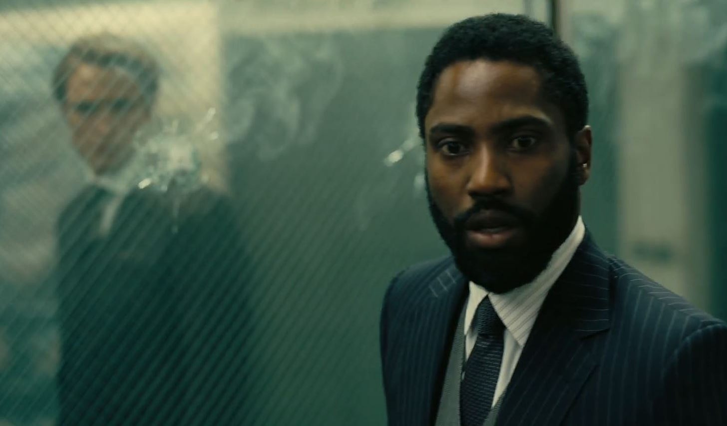 John David Washington in Tenet standing in front of a window with a bullet hole. On the other side of the glass is Robert Pattinson, watching him.