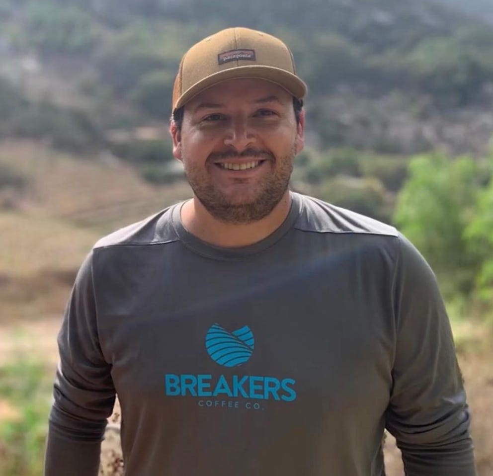 A close-up photo of Kyle Rosa with a blurry agriculture green field behind him. Kyle is wearing a brown hat, has a scruffy beard, and a grey long-sleeve tee with the Breakers Coffee Co. logo on it.