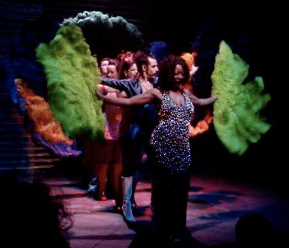 Pictured are five dancers on stage in colorful costumes. They are standing in a row with the author in front wearing a navy dress with silver beading, holding a big green feather fan in each hand. Behind her is the choreographer wearing an electric blue pantsuit and white go-go boots. Each of the dancers holds a set of fans in a different color, which are held at different angles so that the audience can see them all.
