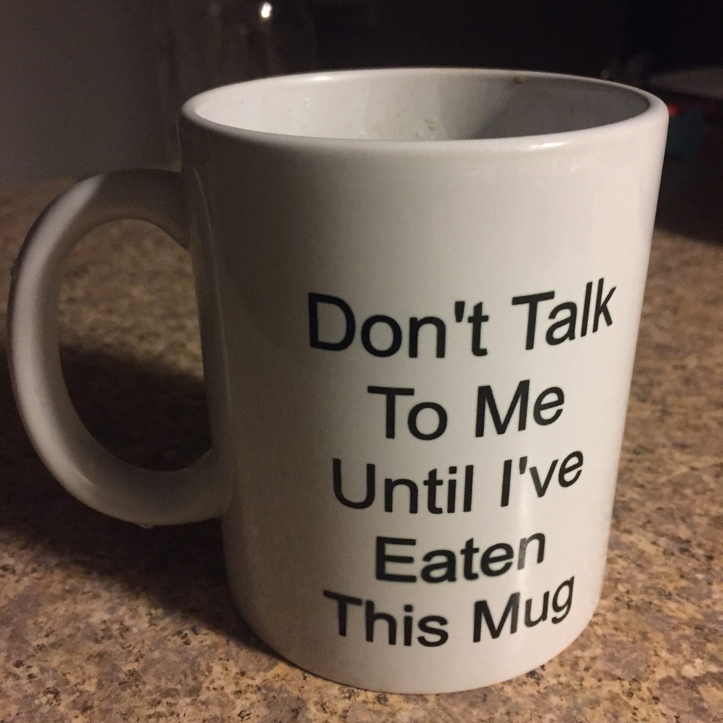 On a counter, a white mug in simple black text that reads "Don't talk to me until I've eaten this mug"