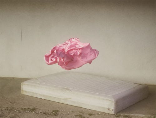 foto montagem. um lençol cor de rosa flutua sobre um colchão ao chão. as paredes são brancas com marcas de uso.