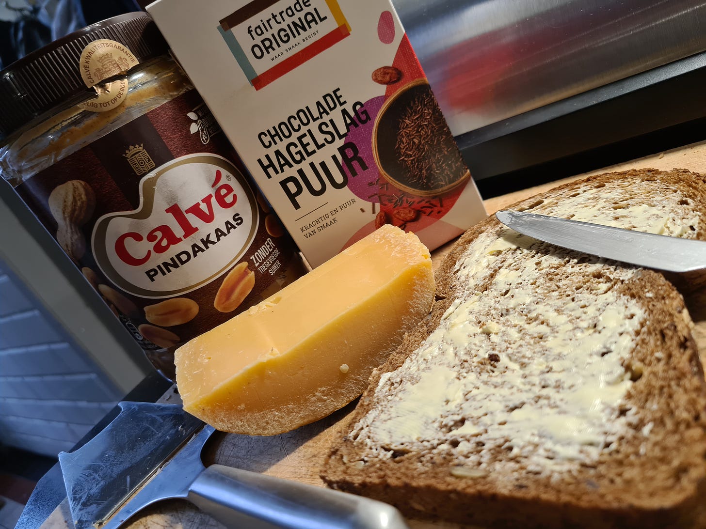 A slice of brown bread, buttered. Behind it, a pice of cheese with a cheese slicer next to it. Behind that, a jar of peanut butter and a box of hagelslag.