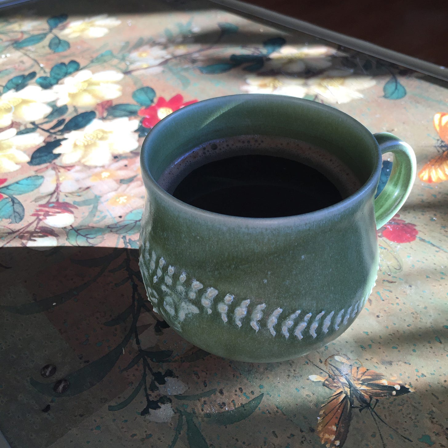 On an old-fashioned tv tray patterned with flours and butterfliers, a teal green mug full of coffee sits in sunlight and casts a shadow to its left. A bone design is embossed on the mug.