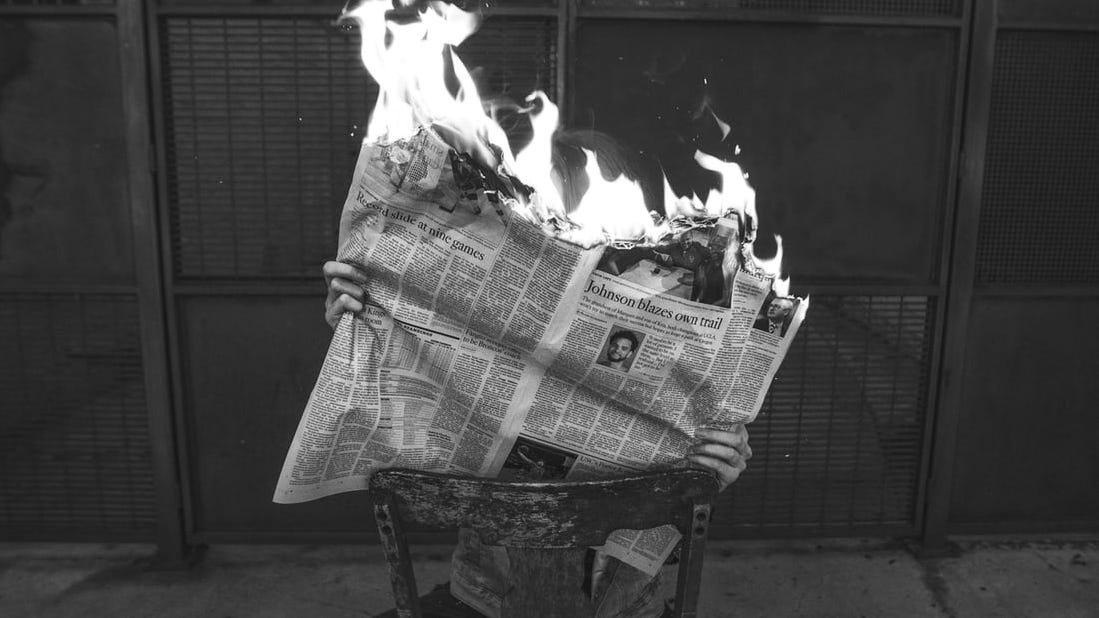 A person sits on a chair reading a burning newspaper