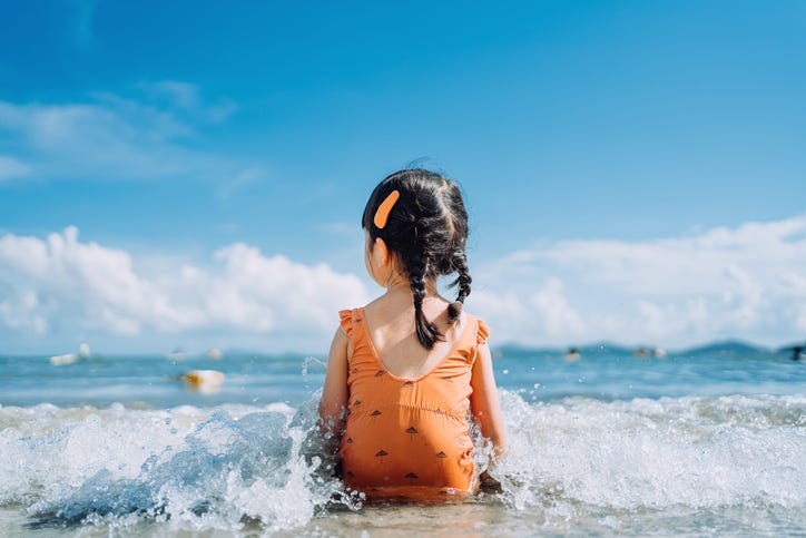 a little girl in an orange swimsuit sits at the edge of the ocean, taking in the view of the horizon as the waves gentle wash over her lap.