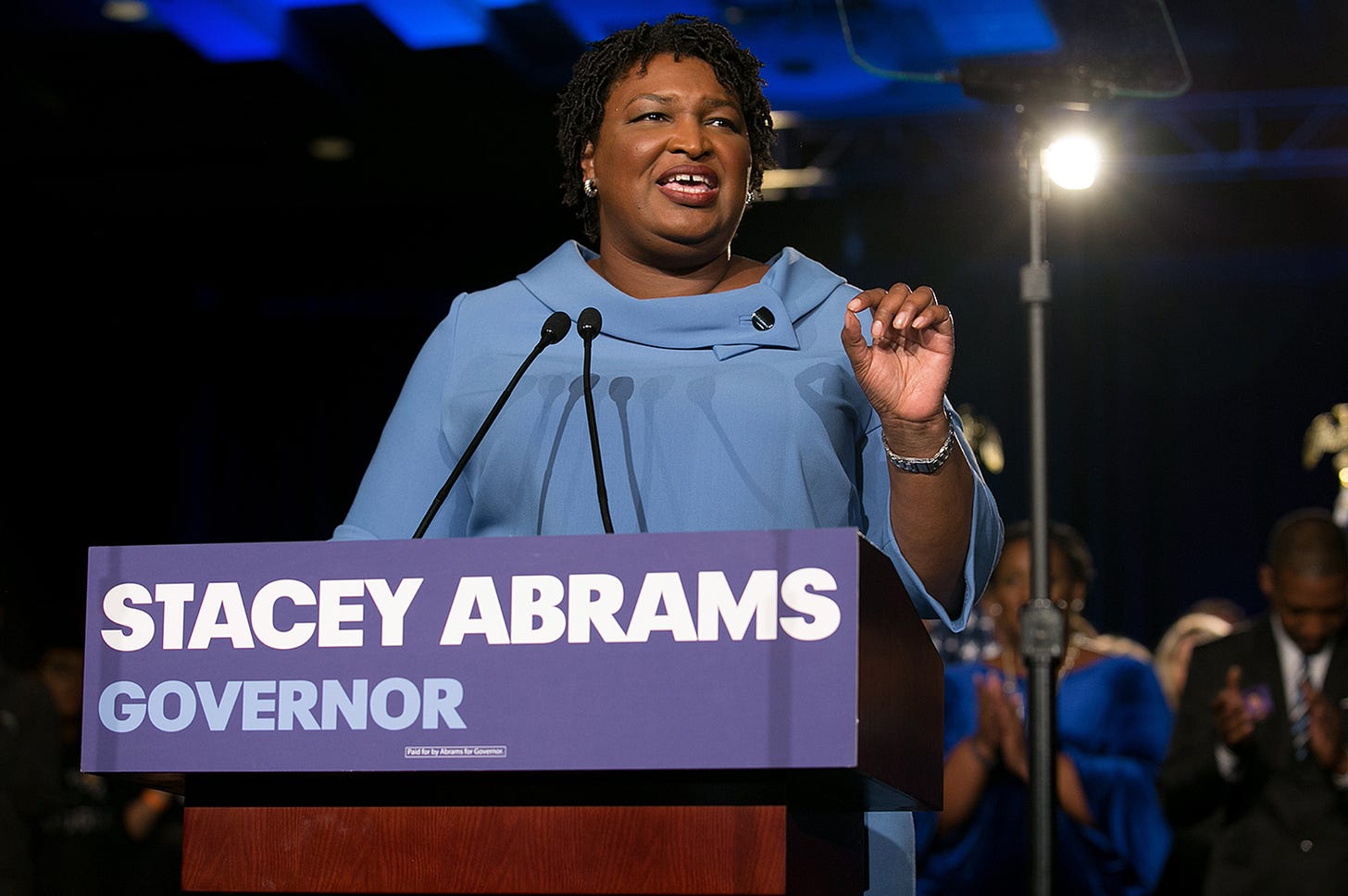 Stacey Abrams is giving a speech during her gubernatorial campaign. She's in a blue dress behind a podium at night.