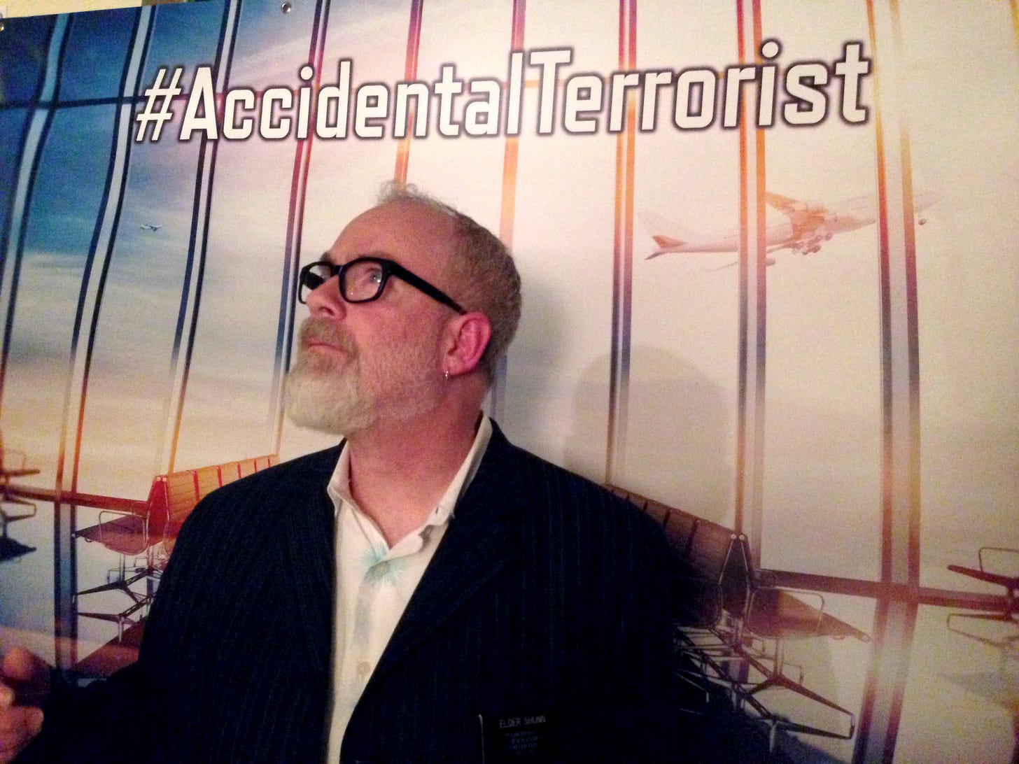A bearded man wearing glasses and a blazer poses before a printed backdrop showing a stylized photograph of the inside of an airport departure terminal, with the hashtag #AccidentalTerrorist printed at the top.