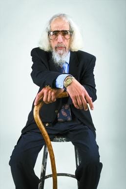 JZ Smith, an older white man, sits on a stool with both hands on a large wooden cane. He has sunglasses on, as well as a dark suit and blue tie. He has long grey hair and a long grey beard.