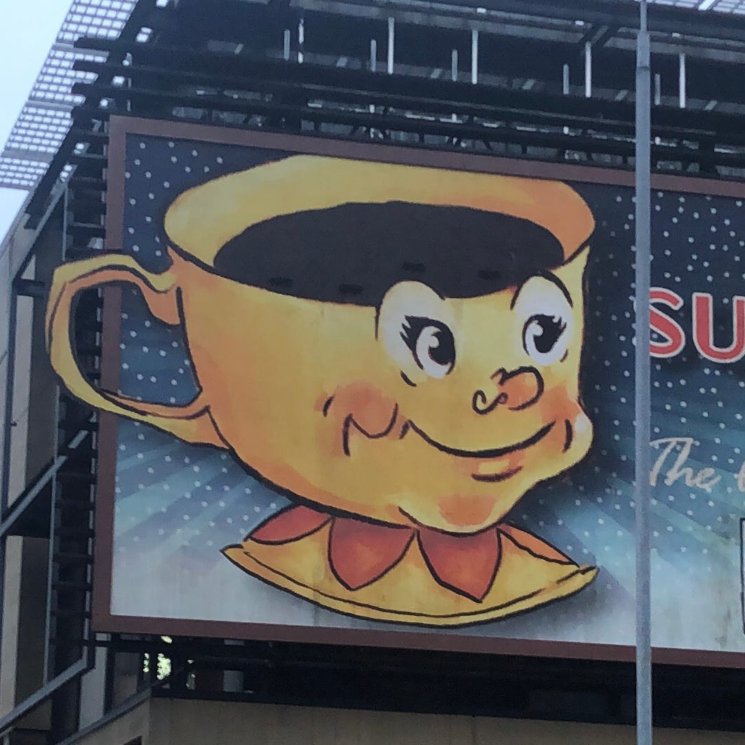 A billboard with an illustration of a personified little cup holding a dark liquid within. It is yellow, with big expressive eyes and a round nose.