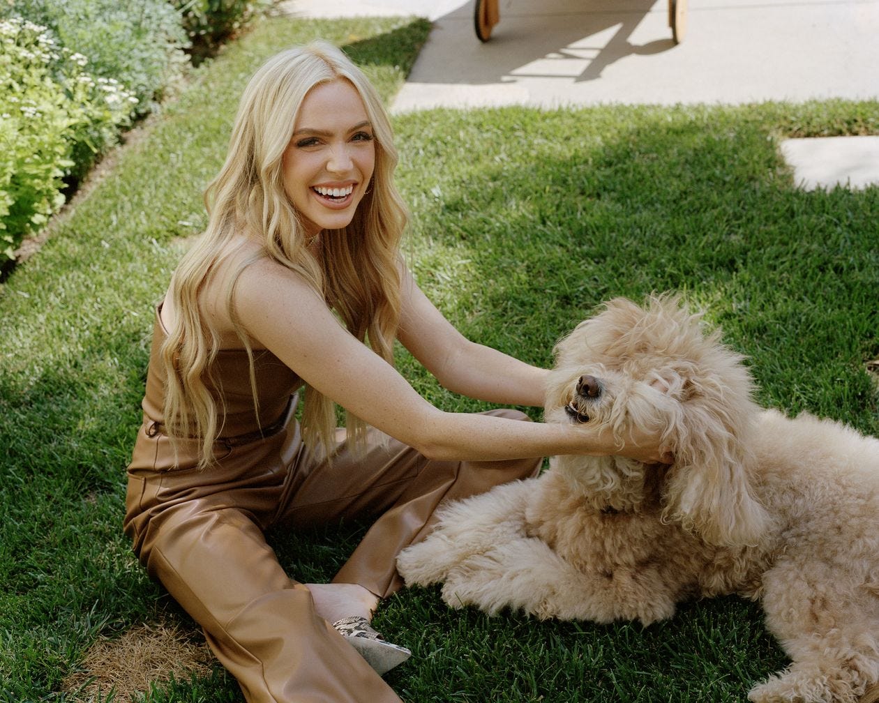 Picture of Alex Cooper, blond and smiling in brown leather pants and a leather tube top (?) sitting on a green lawn and scritching a very fluffy pup.