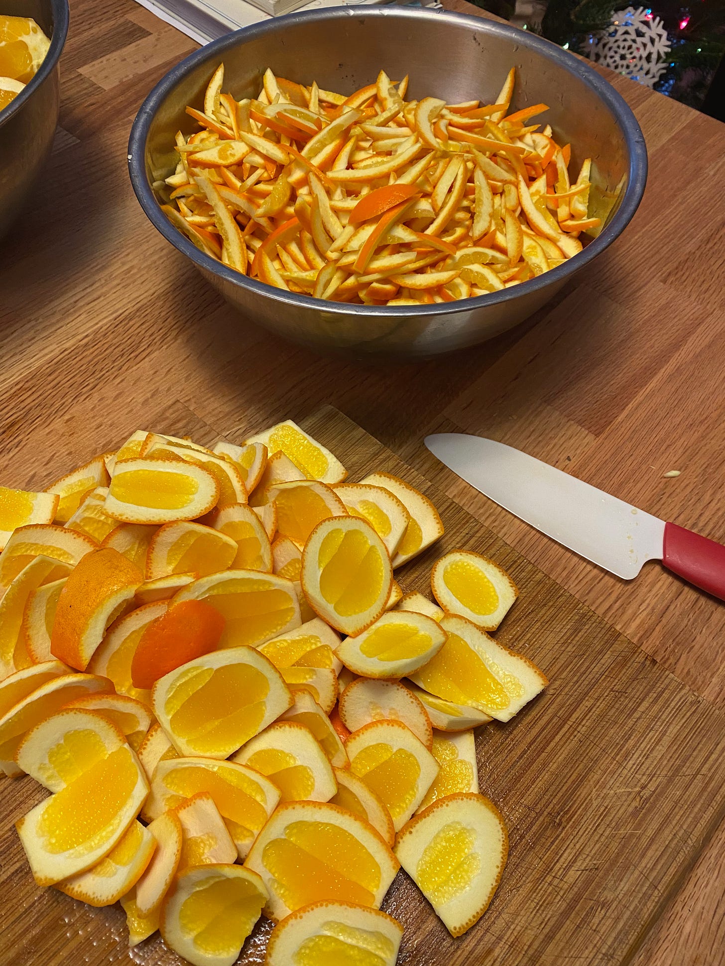 A large silver bowl full of slivered orange peel sits on a wooden counter next to a pile of large pieces of uncut peel.