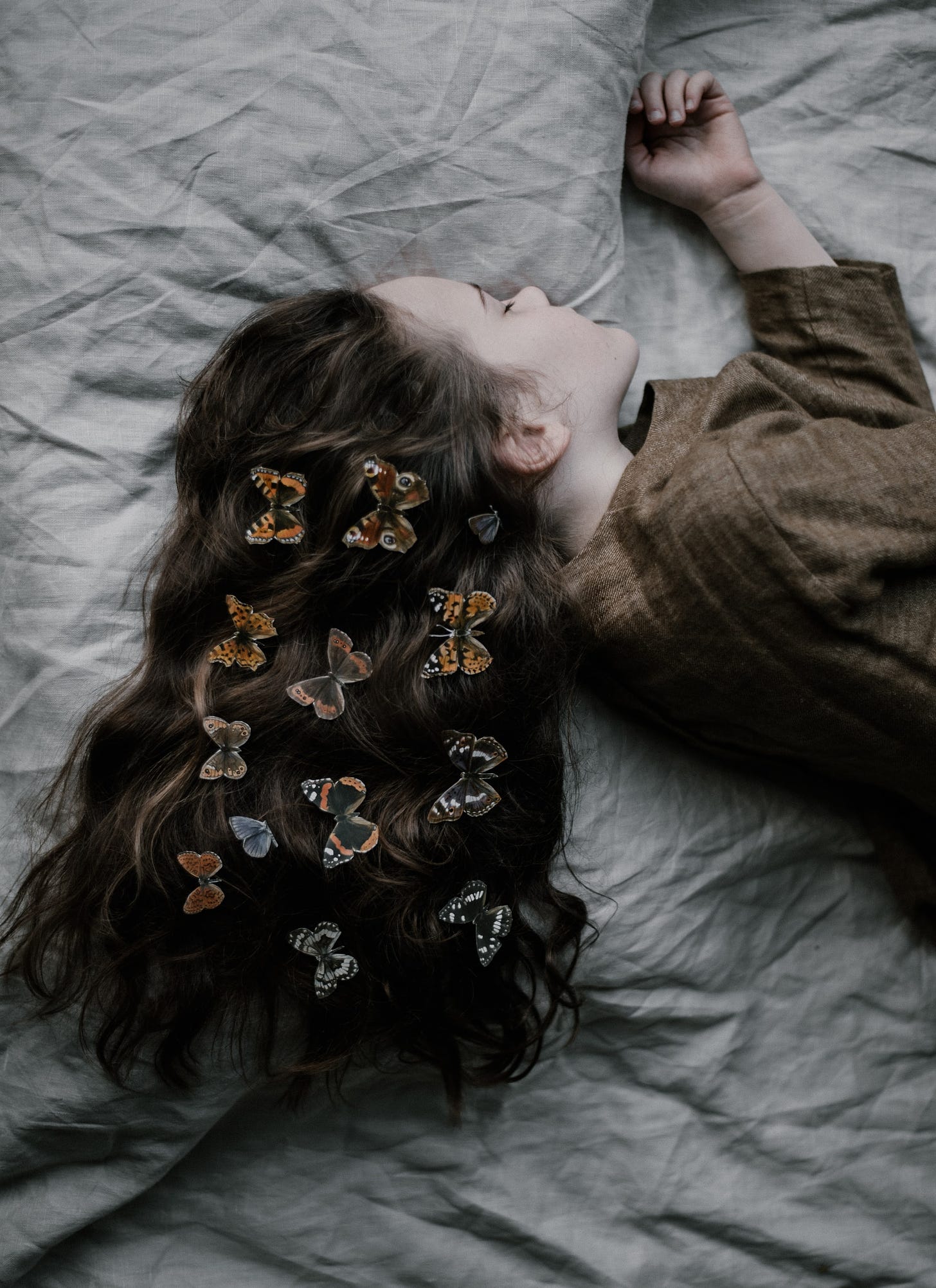 Young girl, laying to the side on a gray blanket. She's asleep, her long dark hair lays spread out behind her. Rows of butterflies of various shade and colors are attached to her hair. She wears a brown dress.