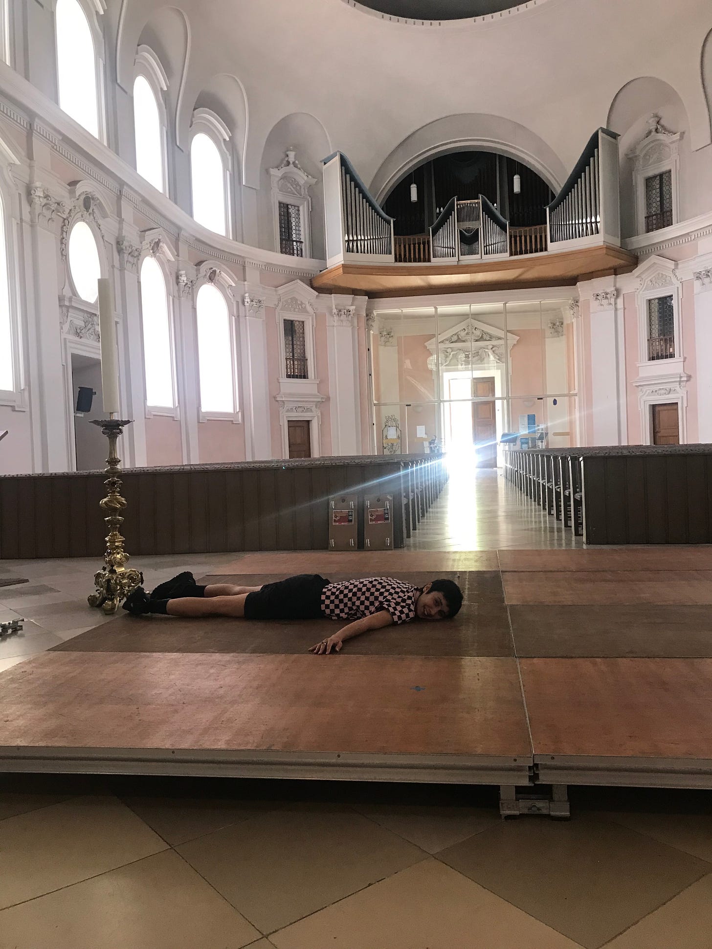 We are inside a big gothic church with pink walls. There is light streaming through the door in the back of the frame. Nikima is lying face down on the dance floor by the altar, smiling and looking at me.