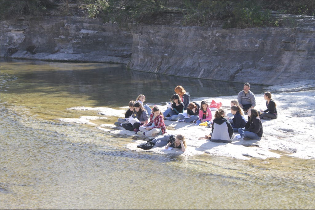 A group of ABC students sit at the edge of a river with journals, reflecting on what to write! 