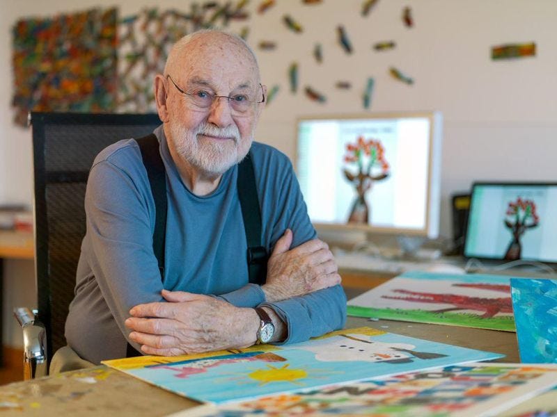 Author and illustrator, Eric Carle, wearing a navy blue longsleeved shirt and black suspenders and clear glasses, sitting at his work desk illustrating with his computer in the background. 