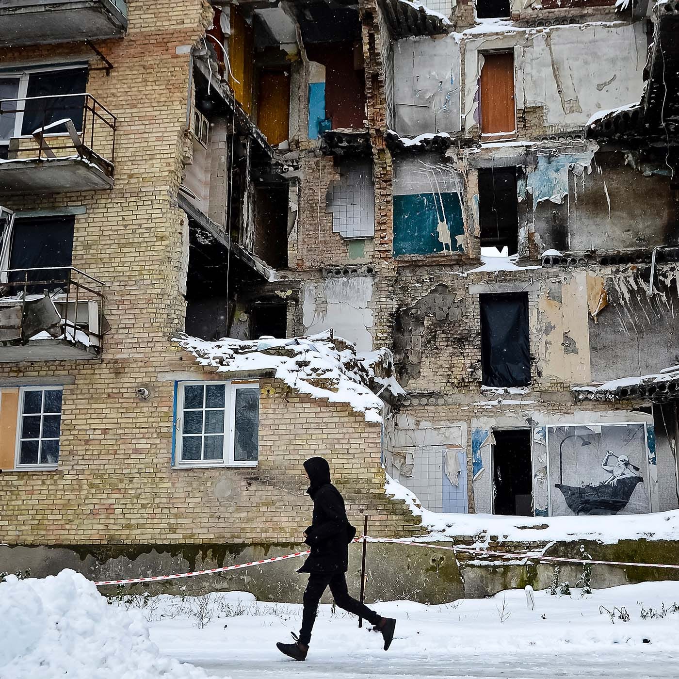 A Ukrainian walks past a bombed-out building. Snow covers everything.