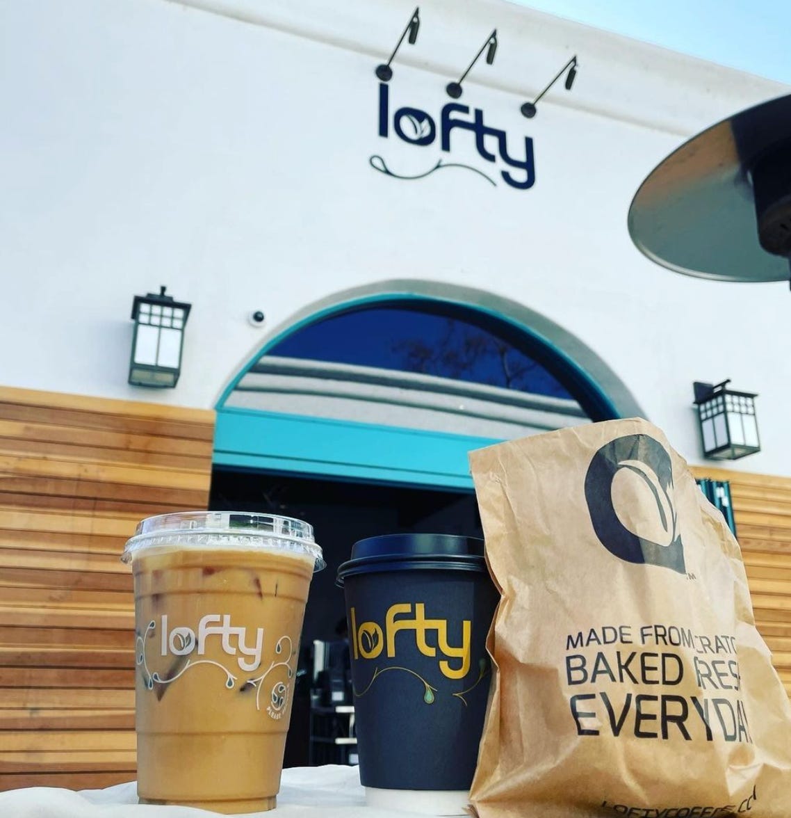 A Lofty Coffee labeled Cold Brew in a clear plastic togo cup, a hot coffee togo and a pastry bag propped up in front of the arched entrance of the Lofty Coffee Shop that has a wood lower wall, white washed top half and the Lofty Logo above the arch.
