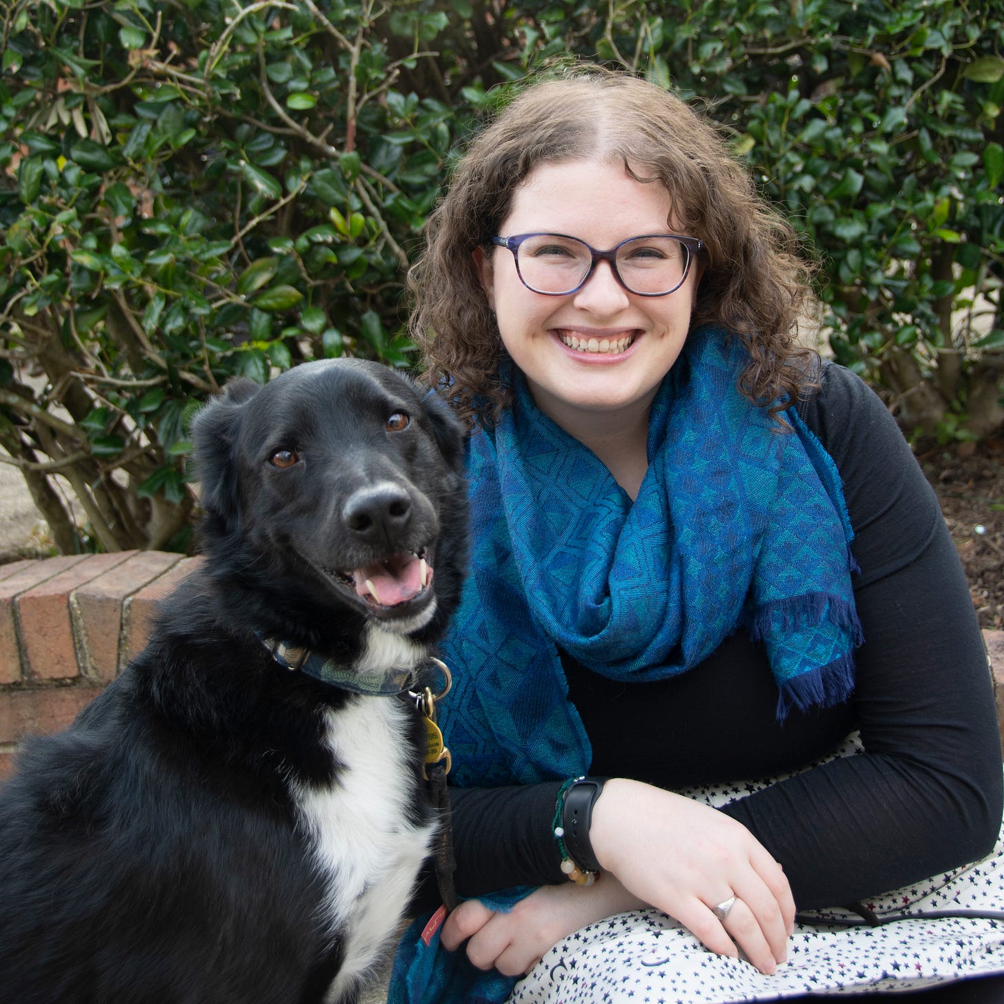 A photo of Elizabeth and her dog Wallace. Wallace, a medium-sized black dog with a white belly, sits on the left with a smile on his face. On the right is Elizabeth, with a blue scarf and purple glasses on, smiling wide on the right. 