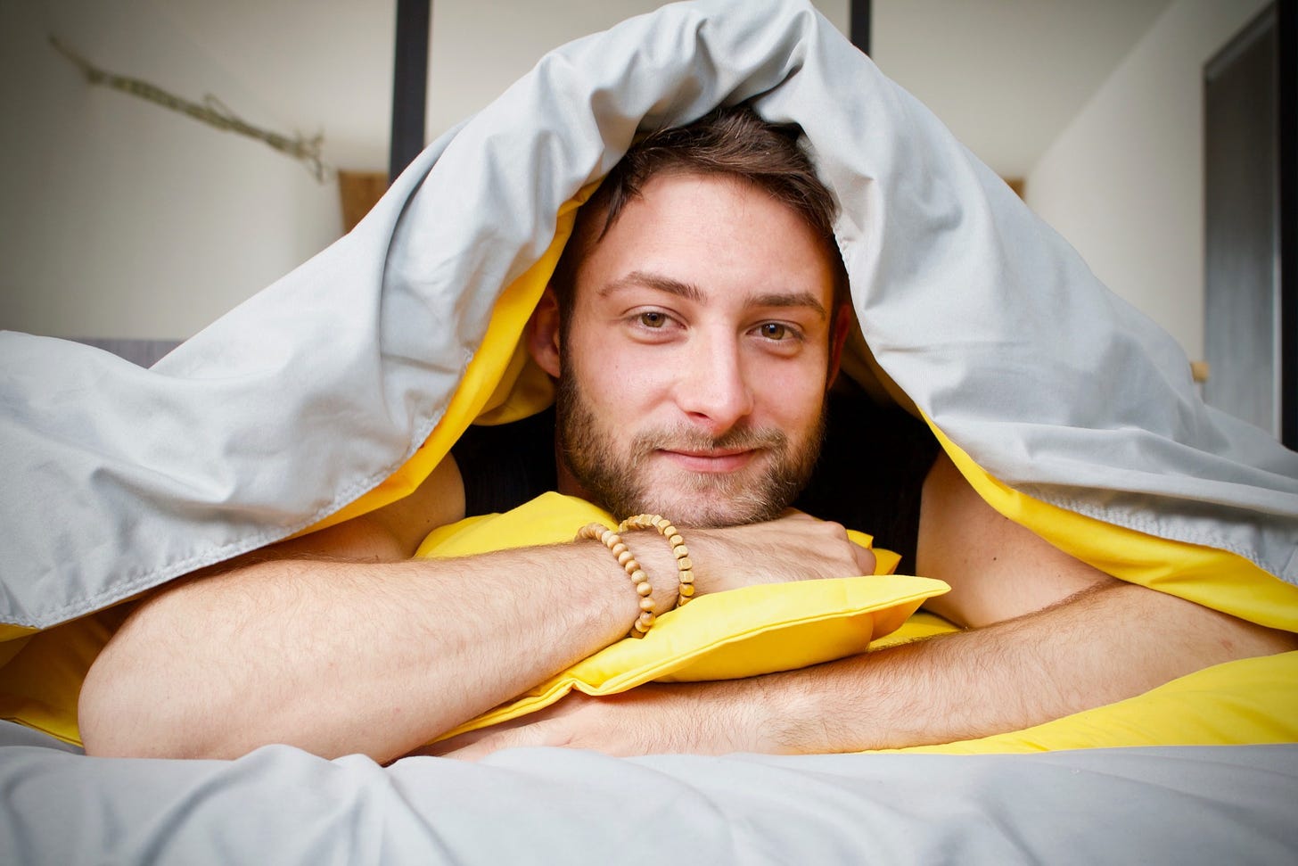 A man lays in bed with his hands folded under his chin, and his blanket over his head. He smiles.
