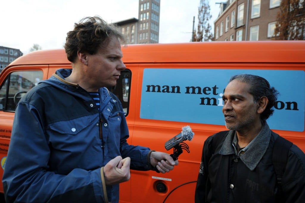 foto van Chris Bajema op het Javaplein in Amsterdam voor zijn oranje Man met de Microfoon bus. Hij interviewt een donkere man.