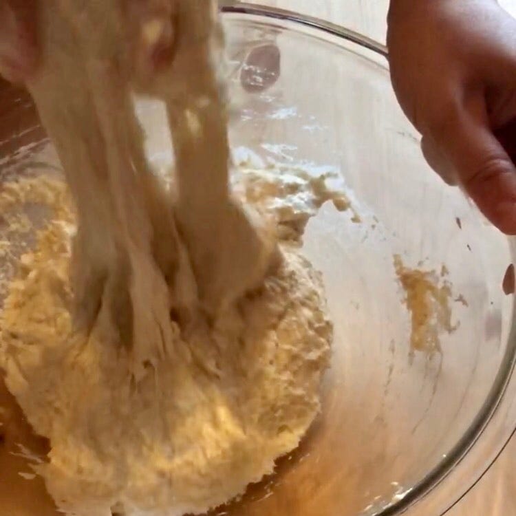 A glass bowl contains dough that is being pulled up by a hand, but the dough is shredding where it is being pulled.