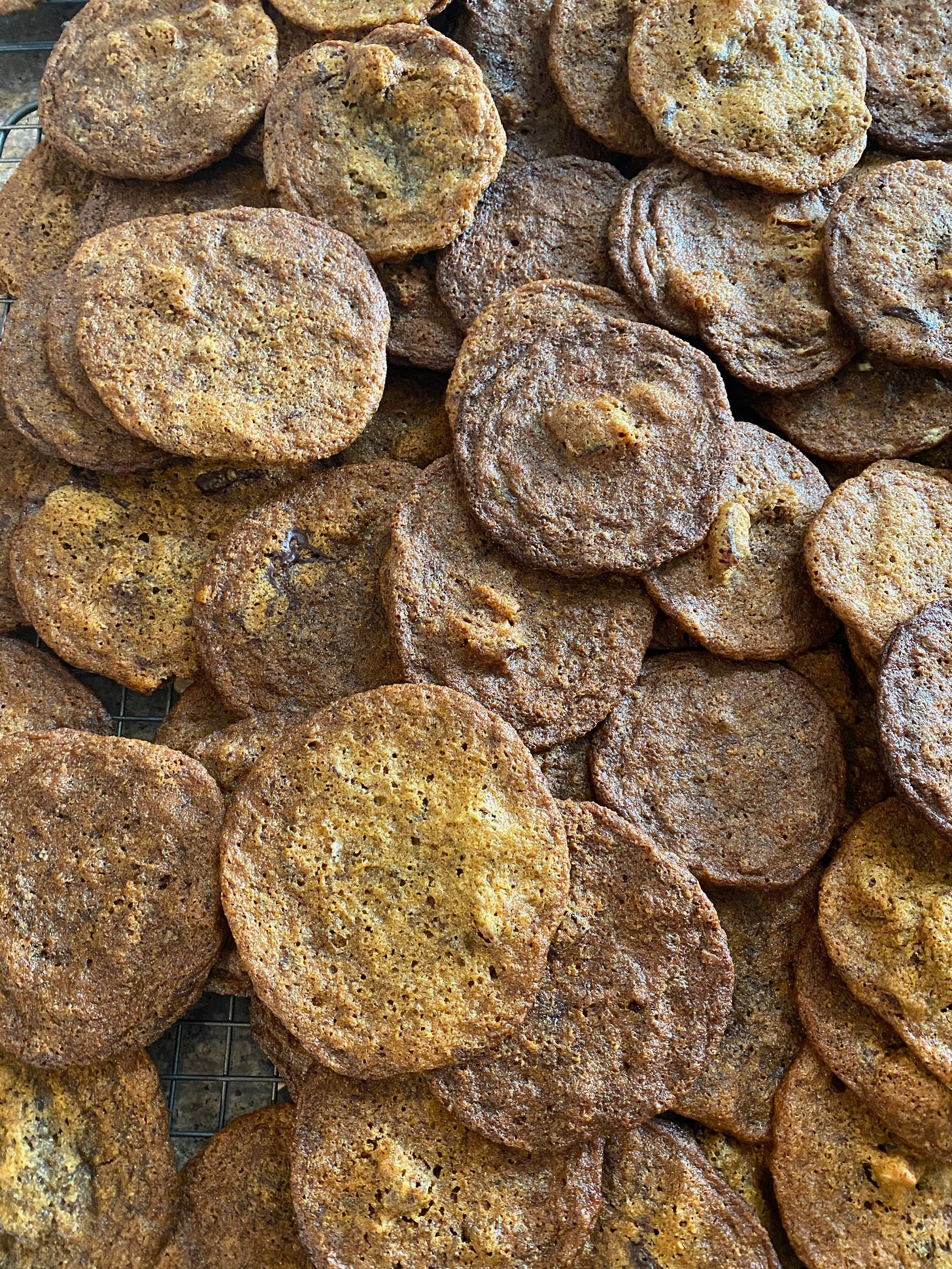 A closeup of many large round cookies piled on top of each other. They are golden brown and crinkly, with some big chunks of chocolate and halvah visible. 