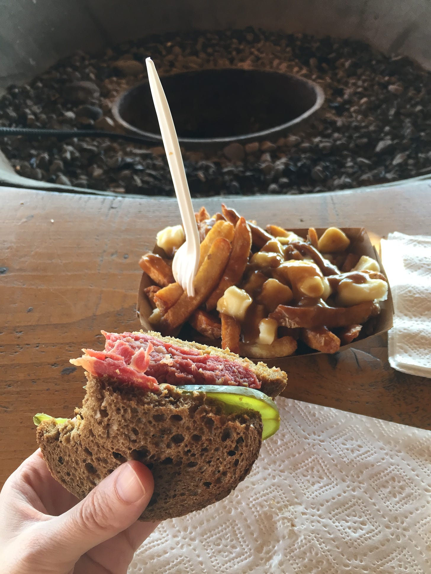 My hand holding a smoked meat sandwich half on rye with one bite out of it. Behind it on a wooden table is a rectangular paper dish of poutine with a white fork sticking out. In the background is a fire pit, unlit.